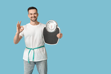 Handsome sporty young man with measuring tape and scales showing OK gesture on blue background....