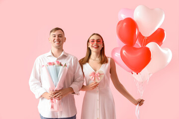 Happy young couple with heart-shaped balloons, bouquet of flowers and gift box on pink background....