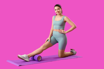 Young woman in sportswear training with foam roller on mat against purple background