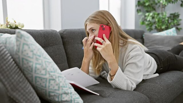 Blonde woman with blue eyes talking on red phone and taking notes at home couch setting.