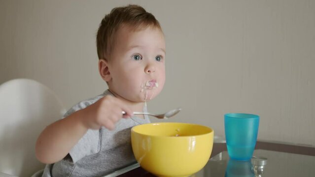 A cute toddler boy eating a healthy breakfast of cereal in the kitchen in the morning. A baby learning to eat with a spoon. Complementary feeding are new stage of feeding of child.