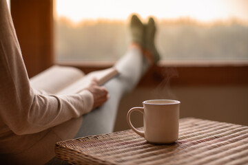 Person relaxing at home reading book feeling relaxed on a cozy winter morning enjoying cup of hot  tea coffee  - Powered by Adobe