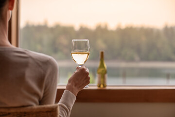 You woman at home relaxing by a window enjoying glass of wine and a beautiful nature sunset lake view 