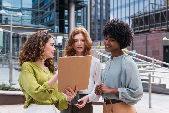 Group of young successful business people chatting outside modern office building and smiling, holding paper