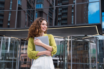 Photo of european woman 20s standing and holding laptop in front of office building