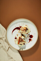 On a decorative plate are two vanilla ice cream balls with almond flakes, cinnamon and chocolate sauce. Brown background with a cloth scarf. Attractive dessert.
