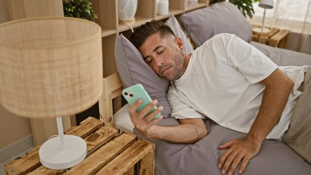 Young, stressed hispanic man lying in comfortable bedroom, engrossed in serious smartphone texting, face showing signs of stress, indoors.