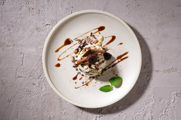 Top view of an ice cream decorated with chocolate sauce and crushed nuts on a white ceramic plate...