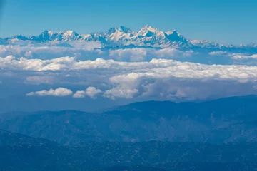 Wall murals Kangchenjunga Aerial view of dusky layers of blue, green, and snow-capped mountain ranges surrounded by cumulus clouds amid clear blue sky captured from the Airplane window.  The mighty peak of the Kangchenjunga.