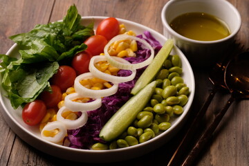 fresh vegetable salad, edamame, sweetcorn, cherry tomatoes, romaine lettuce, sliced cucumber, sliced onions, and purple cabbage. extra virgin olive oil. served on a white ceramic plate. appetizer. 