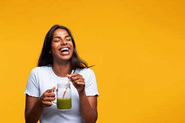 Happy young beautiful Indian woman with green juice on yellow background, laughing. Copy space.