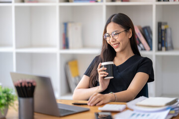 Freelance Asian businessperson, businesswoman sitting and enjoying hot coffee, pay attention on the laptop screen while relaxing.
