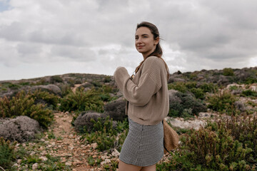 Adorable cute woman with dark hair wearing knitted cardigan is posing against blue sky and...