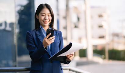 Confident Asian woman with a smile standing holding notepad and tablet at out side office. big city
