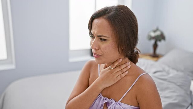 Young, beautiful hispanic woman in pyjamas sitting on her bedroom bed, in visible pain as she suffocates, strangled by unseen health problem.