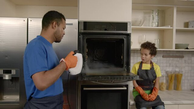 Positive attractive African American father and cheerful cute school age son in protective workwear doing housework together , washing dirty kitchen oven from grease using steam cleaner and detergents