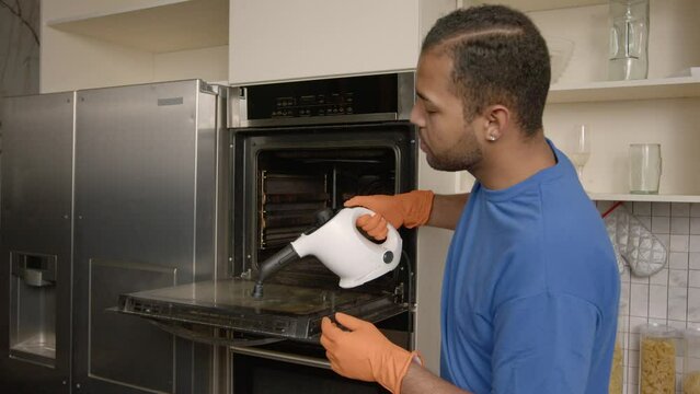 Attractive African American man in protective gloves and apron doing household chores, washing dirty kitchen oven from grease and fat using steam cleaner and detergents in domestic kitchen.
