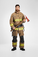 Fototapeta premium Serious male firefighter in uniform holding big axe for work. Front view of dark-haired man in fireman apparel posing with wooden hatchet tool, on gray studio background. Job, equipment, tool concept.