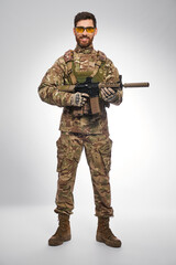 Smiling armed soldier posing with assault rifle, looking camera indoors. Front view of confident...