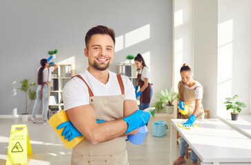 Portrait of happy smiling male young janitor in uniform looking cheerful at camera holding household cleaning supplies in hands. Cleaning team service, housework and housekeeping concept. - Powered by Adobe