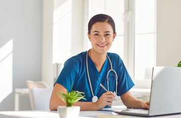 Portrait of cheerful young friendly female doctor in blue medical uniform sitting and working on...