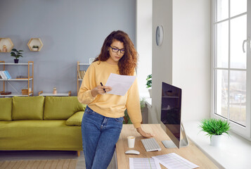 Young serious curly redhead business woman accountant in glasses standing near the desk of her workplace with financial documents with tables at home or at office analyzing company.