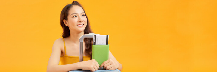 Pretty young lady holding passport and plane tickets, leaning on suitcase over yellow background