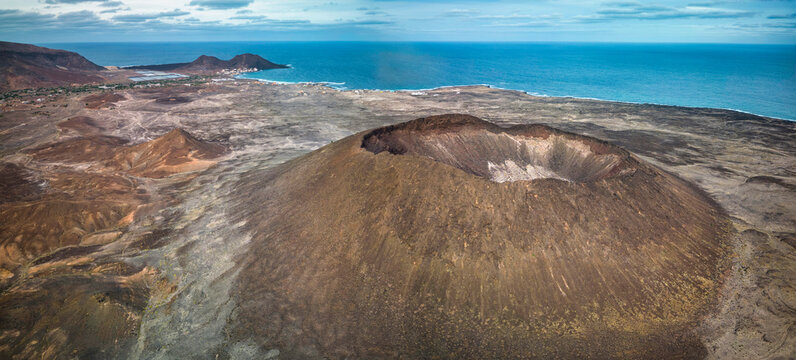 Aerial view of a coastal volcanic landscape (Viana volcano, Sao Vicente) with a distinct crater in the foreground, sparse vegetation, and a small village by the sea, under a cloudy sky
