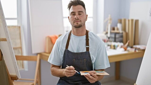 A contemplative young man with a beard wearing an apron holds a paint palette in an art studio.