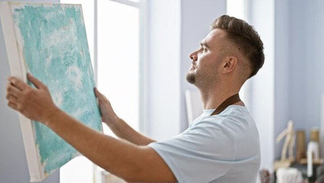 A contemplative man with a beard hangs a blue abstract painting in a bright art studio.