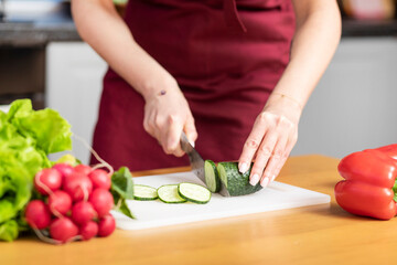 Obraz na płótnie Canvas Housewife in red apron slicing cucumber and preparing vegetarian food 