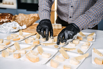 Caterer Preparing Cheese Platters for an Event. Professional caterer arranges various types of cheese on plates, wearing gloves for hygiene while preparing for a gourmet event. - obrazy, fototapety, plakaty