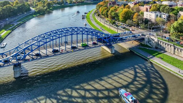 Blue tied-arc Pilsudski bridge over Vistula river in Krakow, Poland. A tram and cars passing the bridge. Tourist ship sailing under it. Flood walls and boulevards on the riverbanks. Aerial 4K video