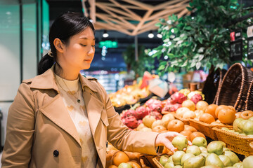 Young Asian woman shopping in supermarket, picking fresh organic vegetables and fruits