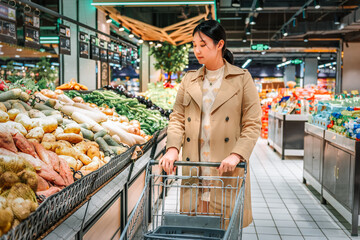 Young Asian woman shopping in supermarket, picking fresh organic vegetables and fruits