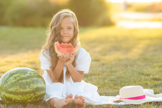 a little girl with long hair in a white summer dress eats a watermelon on the lawn. picnic in the park. happy childhood. space for text. High quality photo