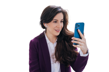 Attractive brunette business woman in violet suit sitting against transparent background using phone makes video call smiles. Cheerful female in headphones with wavy hair makes selfie