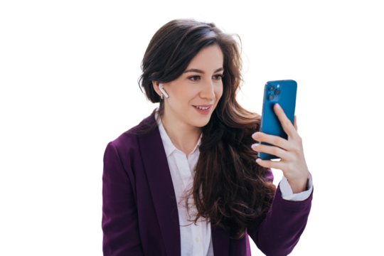 Attractive brunette business woman in violet suit sitting against transparent background using phone makes video call smiles. Cheerful female in headphones with wavy hair makes selfie