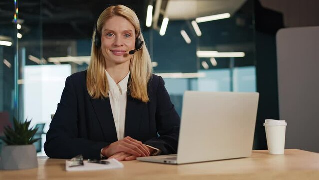 Smiling corporate call center agent with a wireless headset, assisting clients and addressing complaints. Professional telesales woman working in office, looking directly at camera in a webcam view.