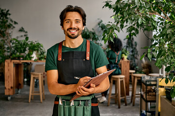 Handsome confident sincere smiling male gardener florist in green t-shirt black apron and bag of garden tools on belt holding wooden clipboard and pen looking at camera. Work in a plant shop. - obrazy, fototapety, plakaty
