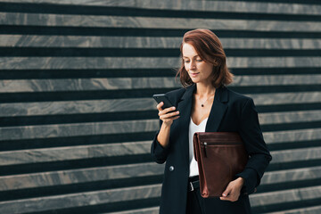 Smiling businesswoman holding a smartphone and leather folder standing outdoors. Middle-aged female with ginger hair typing on smartphone.
