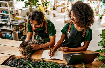 Atmospheric floral boutique. Attractive woman in apron writing in copybook and using laptop while man caressing dark cat at flower shop. Multiethnic family running own small business with love. - obrazy, fototapety, plakaty