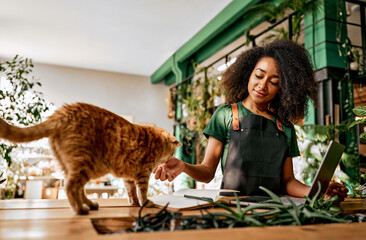 Beautiful confident successful business lady in green t-shirt and black apron petting red cat while working at computer in plant shop. A pet at work with the owner. - obrazy, fototapety, plakaty