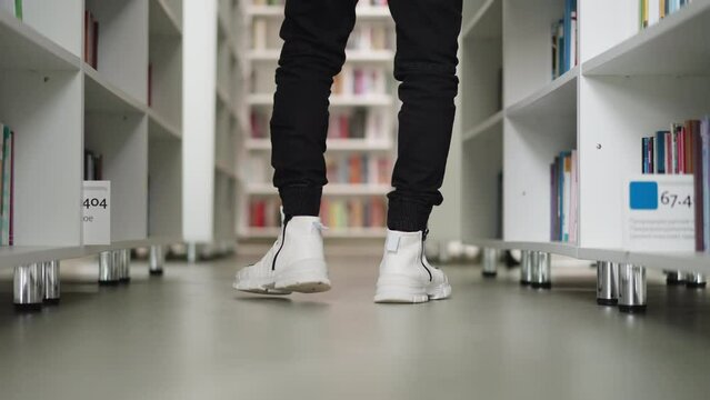 Visitor walks along aisle in library closeup. Man in white boots seeks literature for education on storage racks. Bookstore customer service