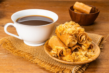 High angle view pile of sweet crispy coconut roll with dried shrodded pork in wooden plate with a cup of hot black coffee on sack cloth on wooden table.
