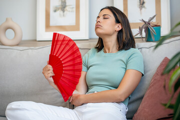 Heated beautiful woman fanning herself while sitting on couch at home.