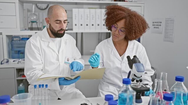 Two dedicated scientists working together, pouring over a document in the chemistry lab, seated indoors at a table with their microscope, engrossed in serious medical research.