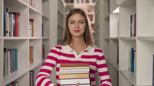Student holds book stack in aisle between bookshelves. Pretty librarian shows literature for customer order in modern bookstore. Smiling lady with folio set