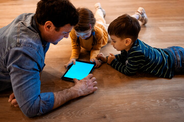 Caring young father with little daughter and son, lying together on floor using digital tablet at home