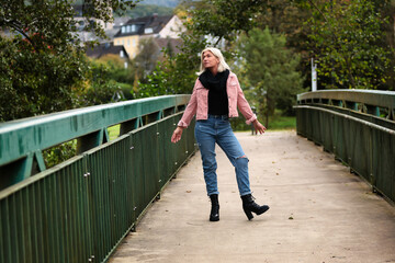 Fototapeta na wymiar Woman blonde in her fifties with a pink denim jacket, blue jeans with a ripped black sweater and a black scarf, portrait on a bridge.